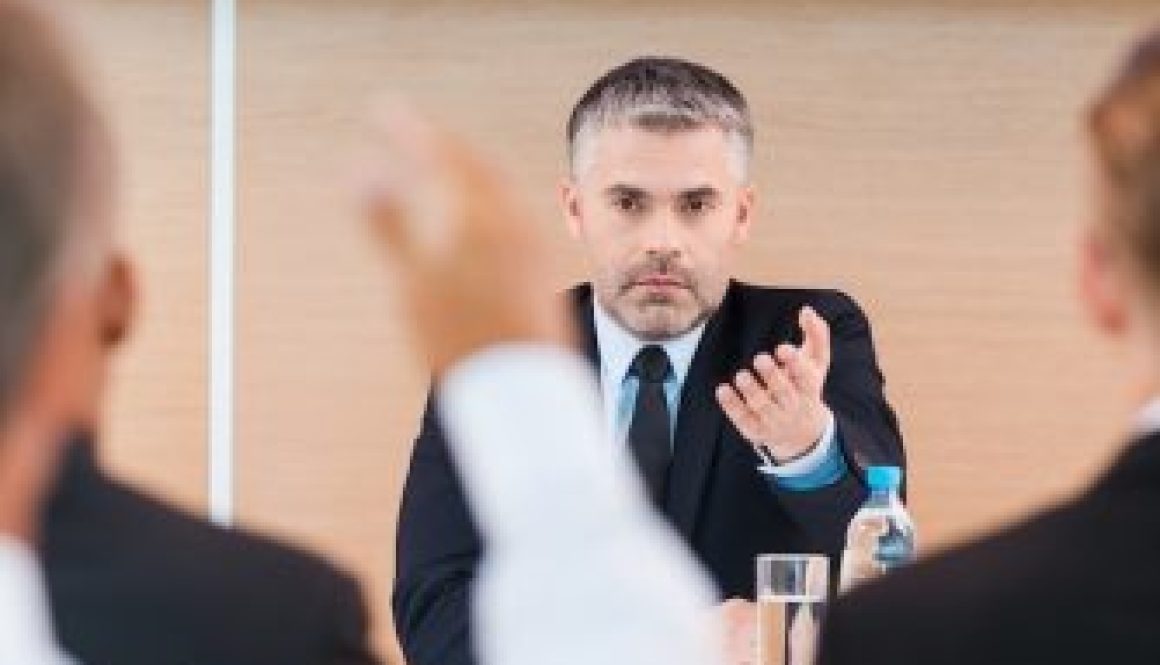 Everyone is welcome for discussion. Confident mature man in formalwear giving a word to someone from audience while sitting in conference hall
